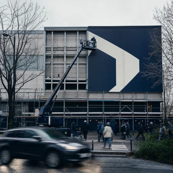 A building facade sign being replaced, a cherry picker raised against half stripped panels above a busy street