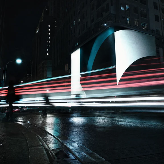 A night street in long exposure, light trails streaking past a large illuminated billboard on wet road